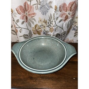 May include: A round, shallow ceramic bowl with a muted sage green glaze and a subtle mottled texture. The bowl features two integrated side handles and a raised circular rim detail, presenting a consistent color throughout the interior and exterior.