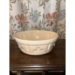 May include: A cream-colored ceramic mixing bowl with a decorative geometric pattern around the rim. The bowl has a rounded shape and a smooth, glossy interior. The bowl is sitting on a dark wooden surface.