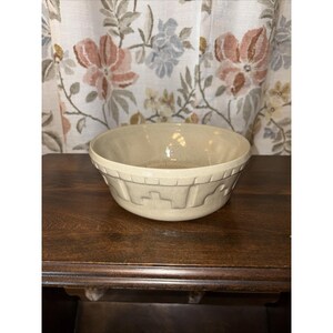 May include: A cream-colored ceramic bowl with a decorative geometric pattern around the rim. The bowl is set on a dark wooden surface, with a floral patterned curtain in the background. The bowl has a slightly aged appearance.