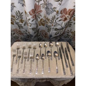 May include: A collection of vintage silverware, including forks, spoons, and knives, arranged on a cream-colored surface. The silverware has ornate detailing on the handles. The background features a floral patterned curtain.