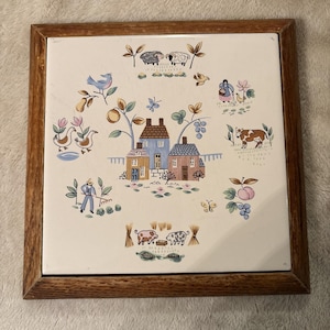 May include: A square ceramic tile framed in wood, featuring a central illustration of three houses surrounded by farm-themed motifs including sheep, a cow, pigs, a person gardening, geese, birds, butterflies, and various fruits and plants.