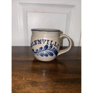 May include: A beige ceramic mug with a blue rim and handle. The mug features the word "GLENVILLE" in blue lettering, along with a blue floral design. The mug is sitting on a dark brown wooden surface.