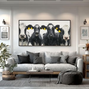 May include: A framed artwork depicting five cows with black and white markings, each with a yellow ear tag. The cows are set against a white backdrop. The artwork is displayed above a grey sofa with dark grey cushions.
