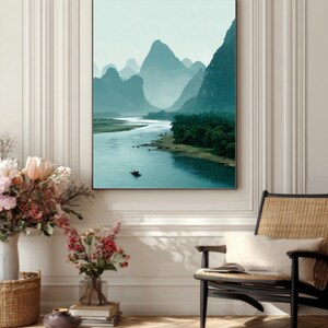 May include: A framed landscape print showing a river winding through teal-toned mountains. A small boat is on the water. The artwork is displayed on a white wall, alongside a woven chair and floral arrangements. The scene evokes a sense of calm.