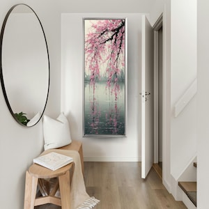 Pode incluir: Uma pintura vertical emoldurada retrata uma paisagem serena com uma cerejeira chorão. Flores rosa caem sobre um corpo de água refletora, com um fundo cinza e verde suave. A obra de arte é exibida em um corredor brilhante.