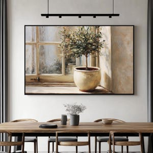 May include: A framed painting depicts a potted olive tree by a window. The artwork features a beige pot with a small tree and green leaves. The window has a white frame and a view of the outside. The painting is in a black frame.