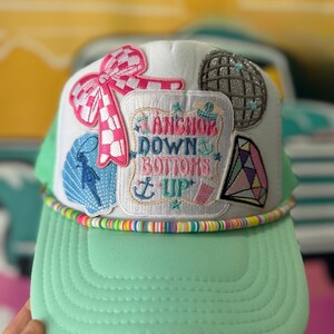 May include: A white trucker hat with a mint green brim and a rainbow-colored trim. The hat features several embroidered patches, including a pink bow, a disco ball, and a patch that says "Anchor Down Bottoms Up".