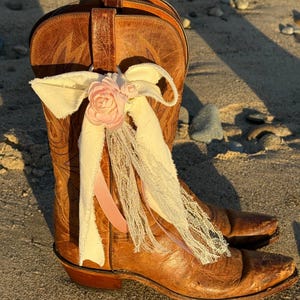 May include: A pair of brown leather cowboy boots decorated with a pink fabric flower and cream-colored ribbons. The boots are worn and have decorative stitching. The boots are on a sandy surface.