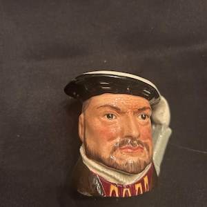 Könnte beinhalten: Keramikfigur eines Männerkopfes mit ernstem Gesichtsausdruck und dunklem Bart. Die Figur trägt einen schwarzen Hut mit weißem Band und einen dunkelbraunen Mantel mit rotem und goldenem Kragen. Der Hintergrund ist dunkel.