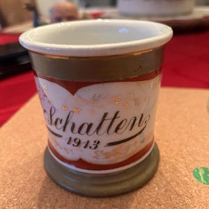 Op de afbeelding: Een kleine, witte keramische beker met gouden rand en een decoratieve rode en gouden band. De beker heeft het woord "Schatten" in elegant schrift, samen met het jaar "1913". De basis is een gedempte groene kleur.