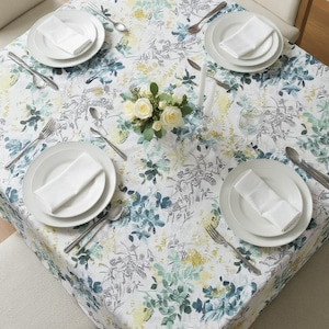 May include: A square tablecloth with a floral pattern in shades of blue, green, and yellow, set for a meal. White plates, napkins, and silverware are arranged around a centerpiece of white roses and candles. The table is surrounded by four beige chairs.