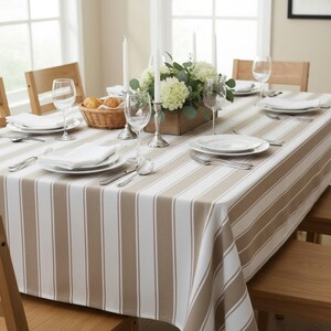 May include: A dining table set for a meal, featuring a striped tablecloth in white and tan. The table is set with white plates, silverware, wine glasses, and white napkins. A wooden box holds flowers and candles.