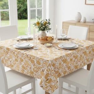 May include: A dining table set for a meal, covered with a tablecloth featuring a repeating pattern of yellow and brown floral designs on a white background. White chairs surround the table, which is set with plates, cutlery, and glassware.