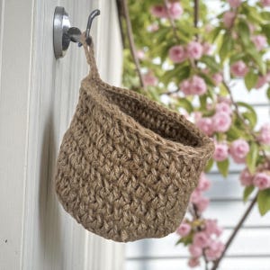 May include: A small, woven basket made of natural brown jute fibre, hanging from a metal hook. The basket has a rounded shape and a loop for hanging. The background features a white wall and pink flowering branches.