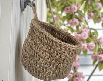May include: A small, woven basket made of natural brown jute fiber, hanging from a metal hook. The basket has a rounded shape and a loop for hanging. The background features a white wall and pink flowering branches.