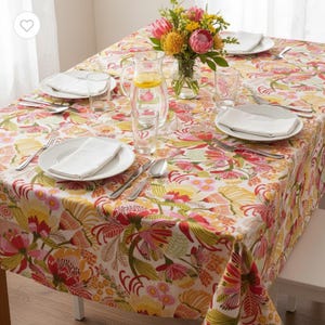 Könnte beinhalten: Ein rechteckiges Tischtuch mit einem lebendigen Blumenmuster in Rot, Gelb und Rosa. Der Tisch ist mit weißen Tellern, Besteck und klaren Gläsern gedeckt. Eine Vase mit Blumen und eine Kanne Wasser mit Zitronenscheiben stehen auf dem Tisch.