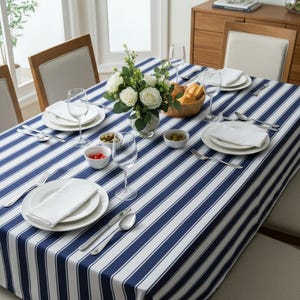 May include: A dining table set for a meal, featuring a blue and white striped tablecloth. White plates, napkins, and silverware are arranged for each place setting. A vase of white roses and small bowls of olives and tomatoes add color.