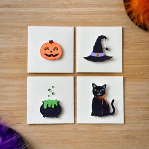 May include: Four square cards with Halloween-themed cutouts. An orange jack-o'-lantern, a black witch's hat with a spider, a black cauldron with green bubbling liquid, and a black cat with an orange bow are displayed.