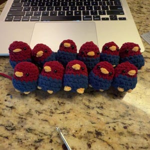 May include: A collection of handmade crochet birds in a row. Each bird has a burgundy head, blue body, and yellow beak and feet. A laptop displaying a screen with the text "but you just have to be strategic" is in the background.