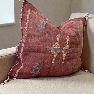 May include: A decorative maroon pillow with embroidered geometric patterns in gold and blue. The pillow is placed on a beige sofa, with tassels on the corners. The pillow has a textured appearance.