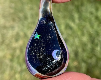 May include: A teardrop-shaped glass pendant with a black background and iridescent glitter. The pendant features a small green star and a crescent moon with iridescent accents.