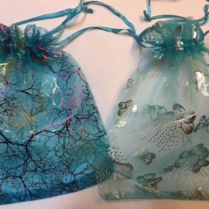 May include: Two turquoise organza gift bags. One features a tree branch design with gold and pink accents. The other has a butterfly pattern with silver details. Both have drawstring closures.