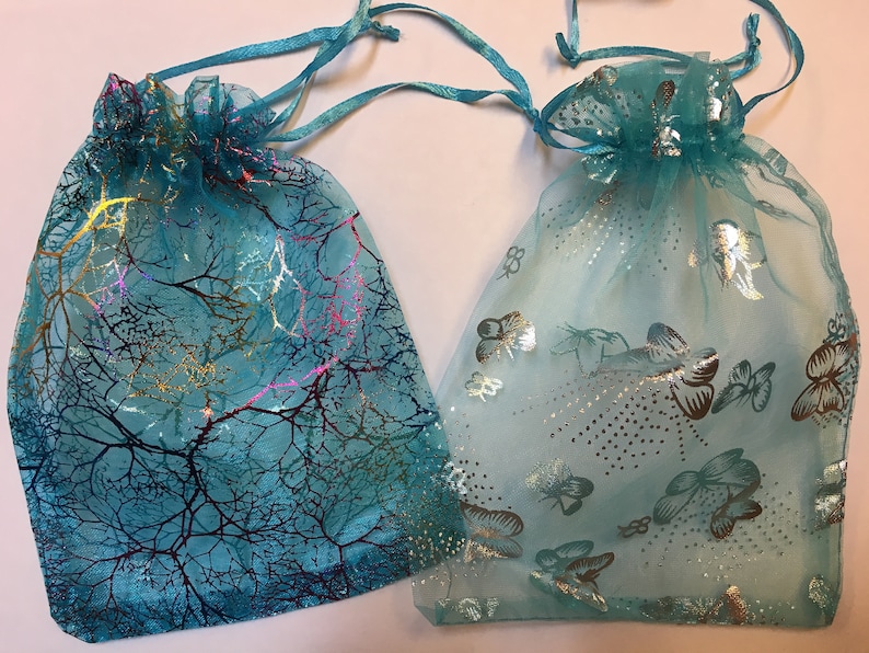 May include: Two turquoise organza gift bags. One features a tree branch design with gold and pink accents. The other has a butterfly pattern with silver details. Both have drawstring closures.
