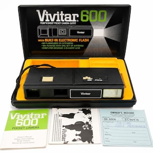 May include: A black Vivitar 600 pocket camera with a built-in electronic flash, housed in a black case with a yellow interior. The set includes an owner's manual, a printed guide, and an owner's record card with the text Vivitar 600 Point'n Shoot.
