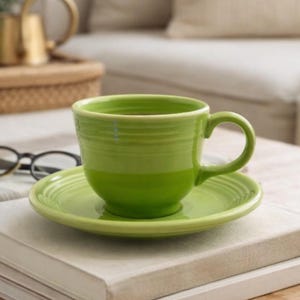 May include: A green ceramic teacup and saucer set. The cup has a rounded shape with a handle, and the saucer is circular. The set is placed on a stack of books, with a blurred background.