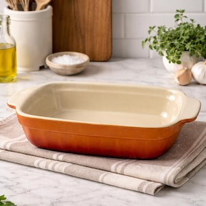 May include: An orange rectangular baking dish with cream-colored interior and handles. The dish sits on a striped beige and white kitchen towel. Other kitchen items in the background include a wooden cutting board, utensils, and a small bowl of salt.