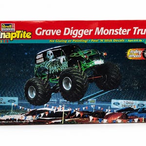 May include: A Revell Monogram SnapTite Grave Digger Monster Truck model kit box. The packaging features a black monster truck with green flame graphics and a skull design, shown jumping over cars in an arena. Text includes Grave Digger Monster Truck.
