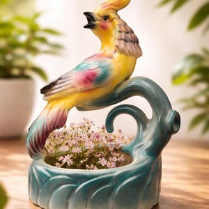 May include: A ceramic bird planter with a yellow, pink, and blue parrot perched on a teal-colored base. The planter holds small pink flowers. The bird has an open beak and a crest. A decorative piece for home decor.