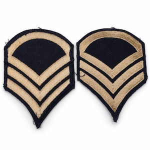 May include: Two matching military-style rank patches featuring a dark navy blue background with embroidered tan chevron stripes and a curved top border. Each patch has a pointed bottom edge and a distinct stitched border around the entire shape.