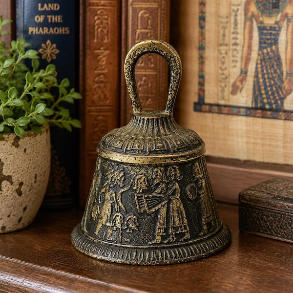 Könnte beinhalten: Eine Metallglocke mit antikem Goldfinish und einem Reliefdesign aus Figuren in historischer Kleidung. Die Glocke hat einen runden Griff an der Oberseite und einen ausgestellten Boden mit dekorativem Rand, der komplexe Prägungen zeigt.
