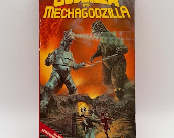 May include: A VHS tape case for Godzilla vs. Mechagodzilla featuring a red and orange background with the title in yellow text. The artwork depicts a battle between Godzilla and Mechagodzilla, with people running in the foreground.