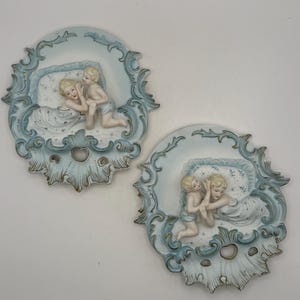 May include: Two oval porcelain wall plaques featuring a relief design of a figure reclining in bed with a small child. The pieces are decorated in shades of light blue and white with ornate, scrolling borders and gold-toned accents.