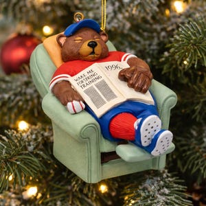 May include: A Christmas tree ornament featuring a brown bear in a red and white shirt, blue shorts, and a blue cap, relaxing in a green chair. The bear is reading a newspaper with the text "WAKE ME FOR SPRING TRAINING" and the year "1996."