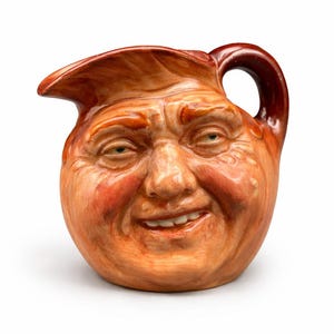May include: A ceramic pitcher with a smiling face design. The pitcher is primarily orange with red accents on the cheeks, nose, and around the eyes. It has a handle and a spout.