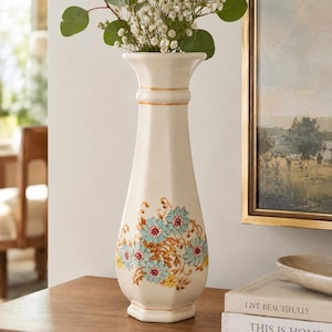 May include: A tall, off-white ceramic vase with a hexagonal base and neck, featuring a painted floral pattern in shades of light blue, yellow, and brown. The vase has a flared rim and contains a bouquet of green eucalyptus and white baby's breath.