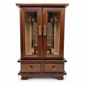 May include: A dark brown wooden jewelry armoire featuring two glass-paneled doors with gold-toned metal hardware and decorative gold filigree patterns. The base includes two small drawers with round metal knobs, and the interior contains hooks.