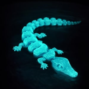 May include: A glowing, articulated alligator figurine in a teal color. The toy is segmented, with a detailed head, four legs, and a long tail. The image is set against a dark background, highlighting the glowing effect.