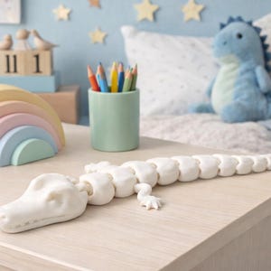 May include: A white, articulated alligator toy lies on a light wood surface. A pastel rainbow stacker, a green pencil holder with colored pencils, and a blue stuffed dinosaur are also visible. The scene is set in a child's room with star decorations.