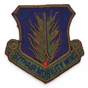 Puede incluir: Un parche bordado en forma de escudo con fondo azul oscuro que presenta un diseño central de tallo de trigo estilizado en verde y marrón. Un pergamino en la parte inferior muestra el texto 97TH AIR MOBILITY WING en letras azul oscuro sobre fondo verde.