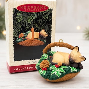 May include: A Siamese cat ornament sleeping in a woven basket filled with green holly leaves, red berries, and a pinecone. The ornament is displayed in front of its original box, which features the text KEEPSAKE ORNAMENT and Cat Naps.