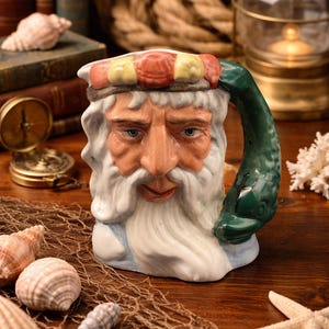 May include: A ceramic mug shaped like an old sailor's face, with a white beard and a hat with red and yellow stripes. The handle is a green fish. Seashells, a compass, and a lantern are in the background.