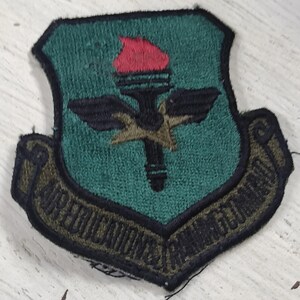 Op de afbeelding: Een schildvormige geborduurde patch met een groenblauwe achtergrond met een zwarte fakkel met een rode vlam, zwarte vleugels en een gouden ster. Een gebogen banner onderaan bevat de zwarte geborduurde tekst AIR EDUCATION AND TRAINING COMMAND.