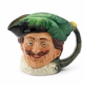 May include: A ceramic pitcher shaped like a man's head, featuring a green feathered hat and a black brim. The face has a mustache and a white ruffled collar. The handle is dark green.