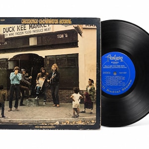 May include: A vinyl record and its square cardboard sleeve for the album Willy and the Poor Boys by Creedence Clearwater Revival. The sleeve features a photograph of a band playing music in front of a storefront labeled Duck Kee Market.