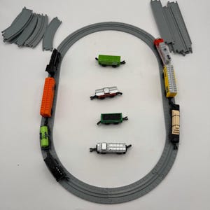 May include: A toy train set featuring a gray oval track, a locomotive, and several colorful freight cars including orange, green, black, yellow, and silver units. Additional loose track pieces and four separate train cars are arranged inside the loop.