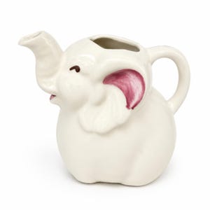 May include: A white ceramic pitcher shaped like an elephant with a trunk serving as the spout and a handle on the back. The elephant features painted dark brown eyes and pink accents on the inner ears, with a smooth, glossy finish throughout.